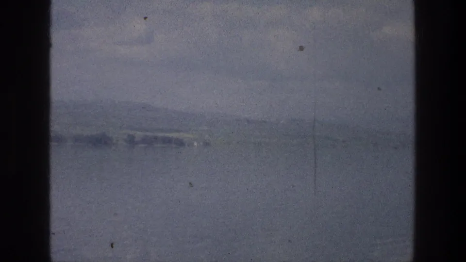 1976 8mm view of the Sea of Galilee with calm water, haze, distant low hills, and dark film-frame edges.
