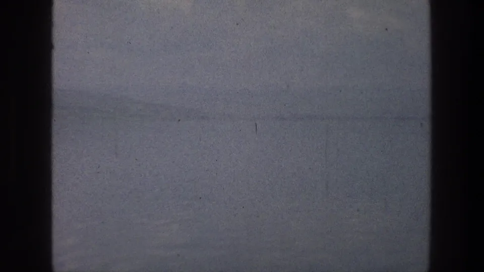 Muted 1976 home-movie frame of the Sea of Galilee with calm water, faint horizon, film grain, and a dark vertical frame edge.