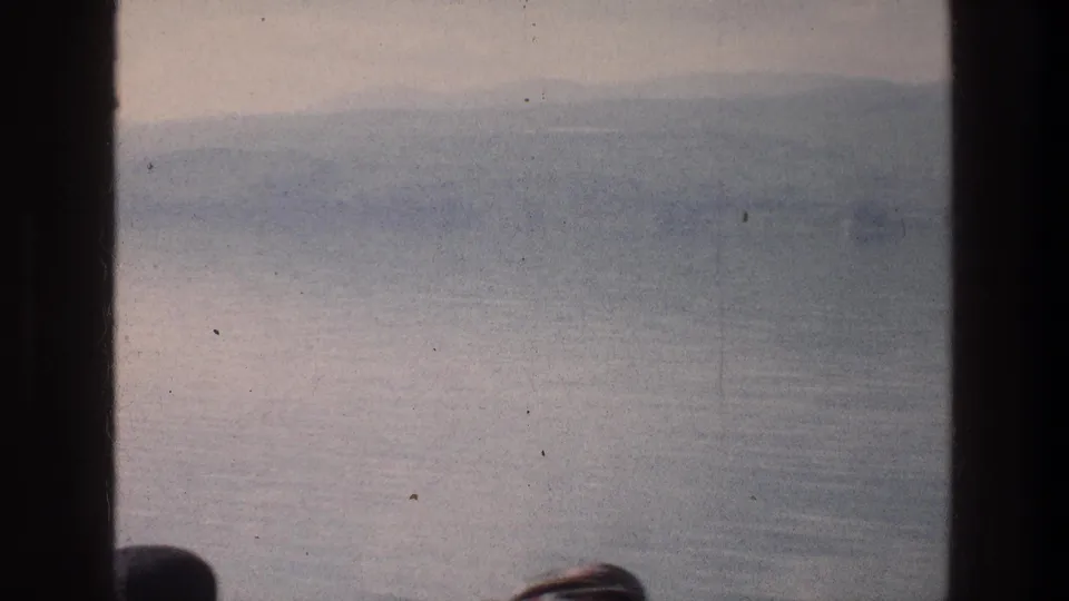 A 1976 home-movie view of the Sea of Galilee shows calm water, distant hills, window framing, and partial passenger silhouettes.