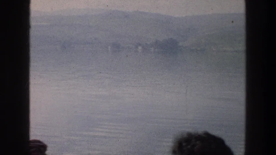 1976 8mm view across the Sea of Galilee in Israel, framed by boat window edges and partial passenger heads.