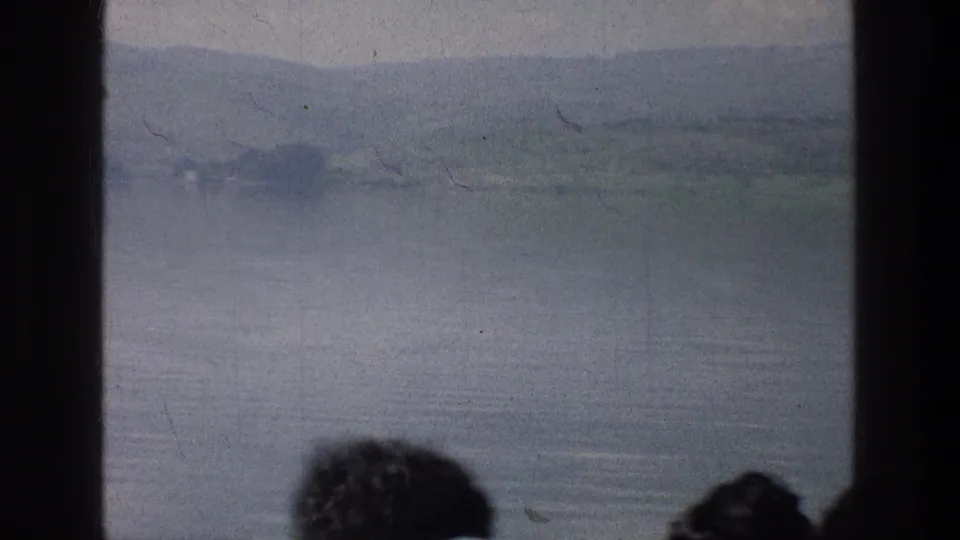 Sea of Galilee landscape in 1976 home-movie footage, viewed past dark window dividers and cropped passenger heads.