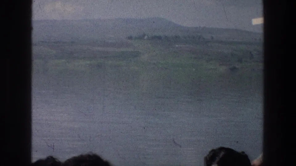 Sea of Galilee boat-view frame from 1976 8mm footage, with hazy hills, water, window bars, and partial passengers.