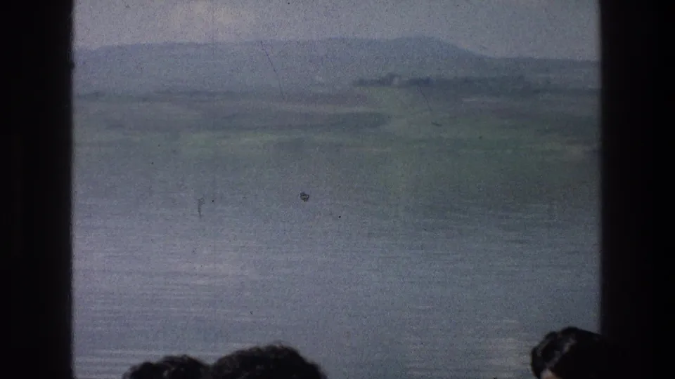 1976 8mm view of the Sea of Galilee with hazy hills, calm water, dark foreground dividers, and partial heads at the bottom.