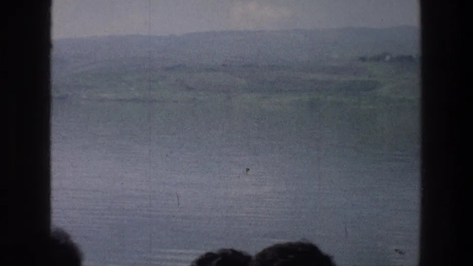 Sea of Galilee landscape in soft 8mm color, seen through a dark central divider with low hills and partial heads near the bottom edge.