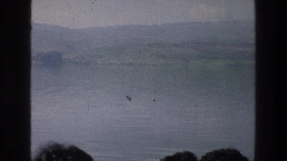 Soft 1970s 8mm lake view at the Sea of Galilee, with muted hills, calm water, dark framing bars, and partial viewer silhouettes.
