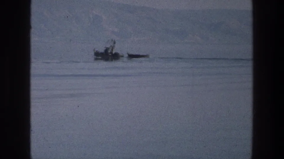 A small boat appears on the Sea of Galilee in 1976 home-movie footage, with hazy hills beyond and partial heads along the bottom frame.