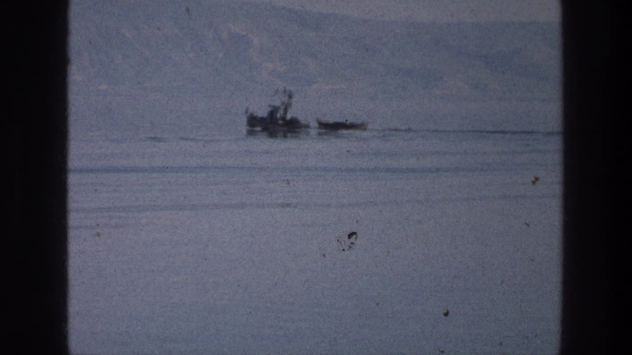 Small boat and narrow craft on calm water with hazy hills in the background.