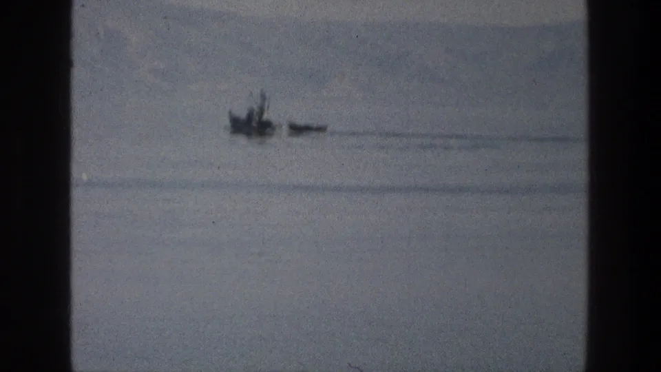 1976 8mm Sea of Galilee frame showing a small dark craft-like shape crossing calm hazy water near a soft distant ridge.