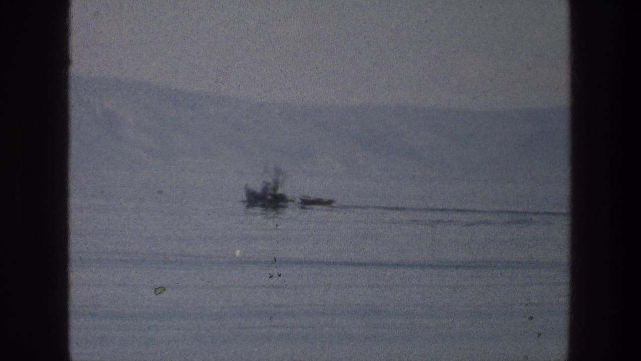 Small dark craft and wake on calm water beneath a hazy shoreline ridge.