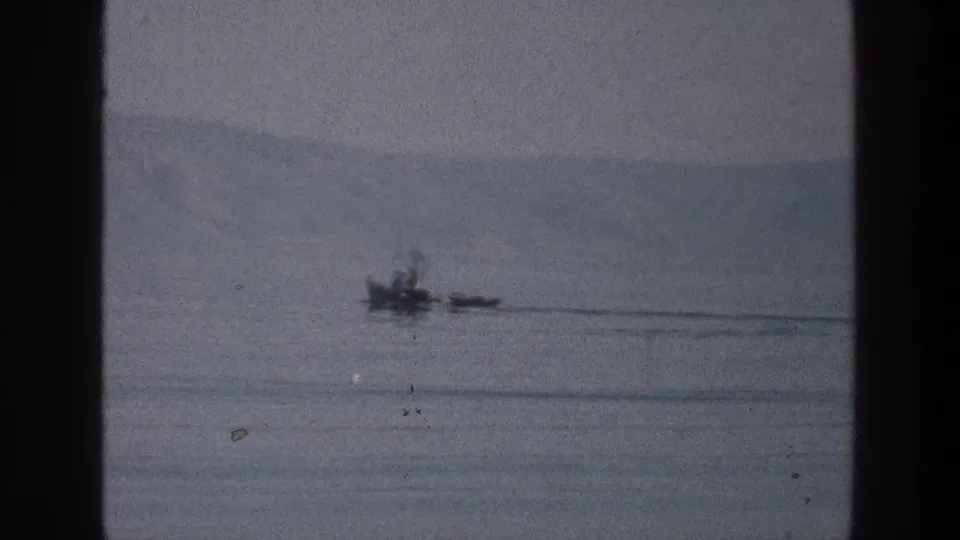 1976 8mm view of the Sea of Galilee with a small craft-like form and trailing wake against a muted shoreline.