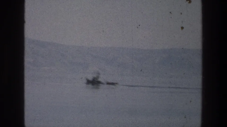 1976 Sea of Galilee 8mm frame with a small dark moving object, faint wake, and distant hazy hills across the water.