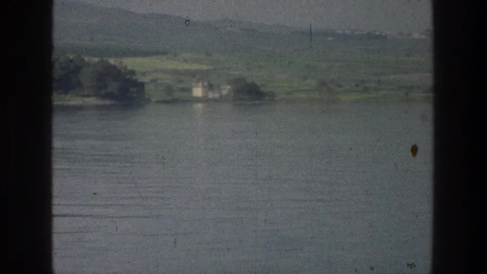 Sea of Galilee shoreline in 1976 8mm footage, showing calm water, distant trees, low buildings, and hazy hills.