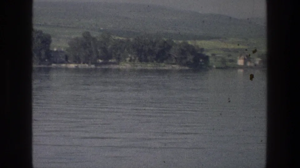 1976 8mm Sea of Galilee view with smooth lake water, a tree-lined shore, low hills, haze, and visible film gate border.