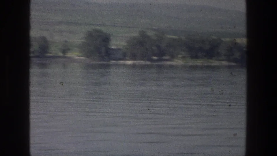 1976 8mm view of the Sea of Galilee with rippled water, a tree-lined far shore, and hazy green hills beyond.