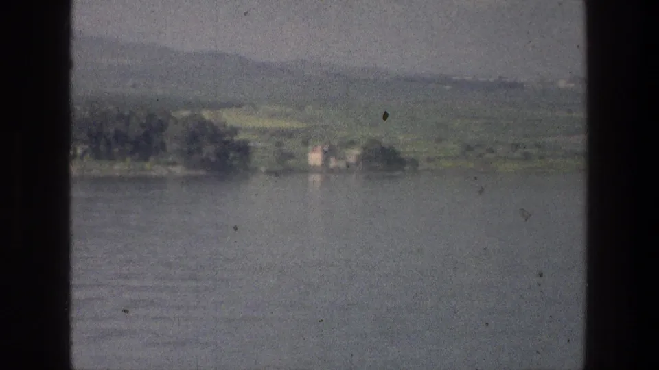 Sea of Galilee view in 1976 showing calm water, a distant tree-lined shore, hills, and a small white lakeside building.
