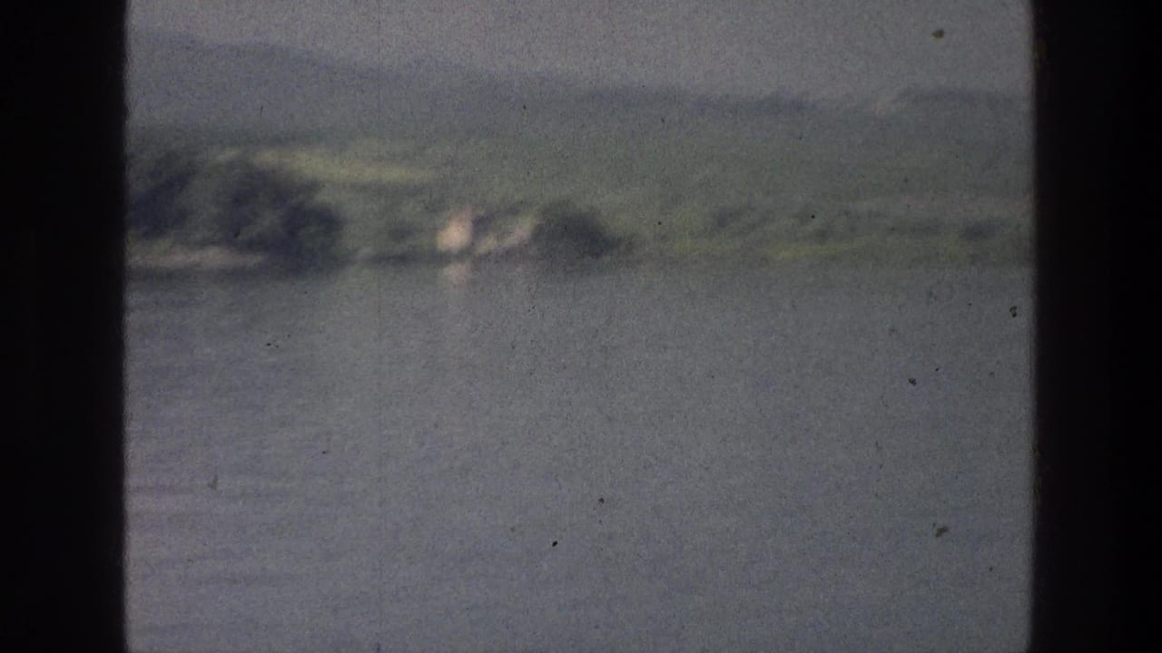 1976 8mm Sea of Galilee view from a boat, with calm water, distant green shore, low hills, and dark window framing.