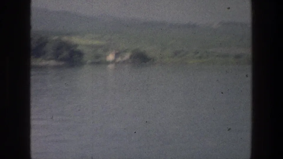 1976 8mm Sea of Galilee view from a boat, with calm water, distant green shore, low hills, and dark window framing.