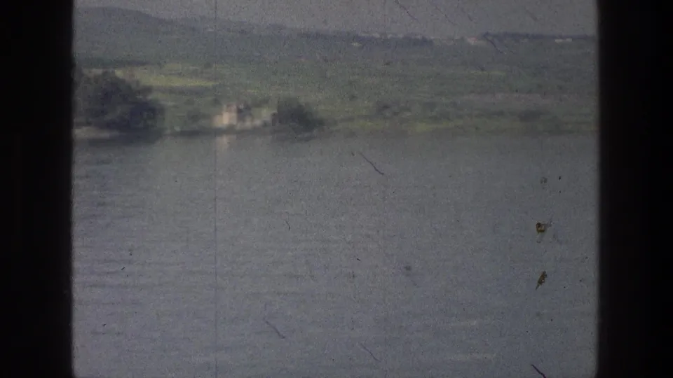 Sea of Galilee shoreline seen from a moving boat, with calm water, green land, and a small pale building in the distance.