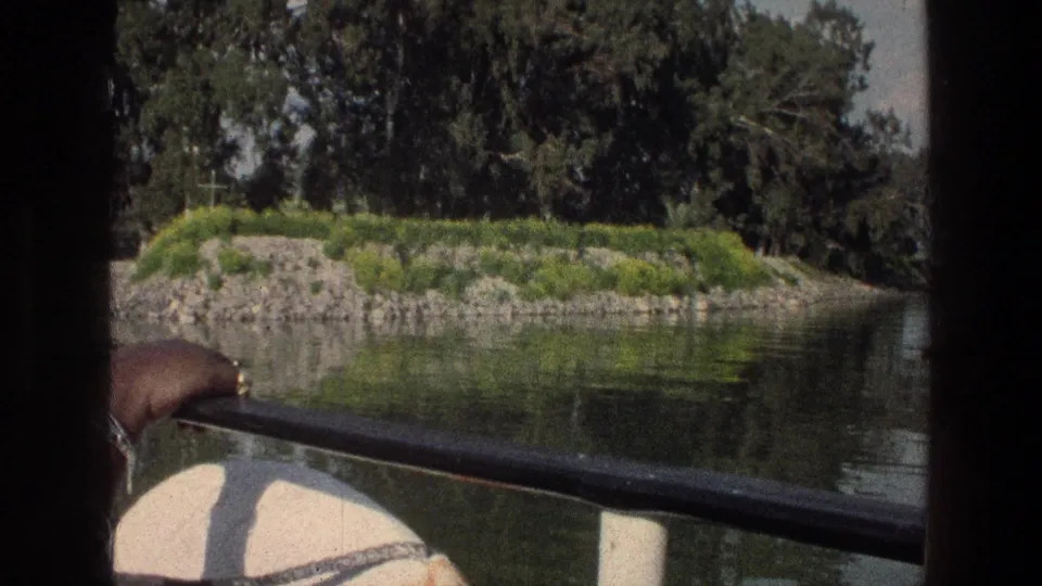 Closer 1976 boat view near a rocky grassy islet on the Sea of Galilee, with trees, reflections, rail, and a passenger hand at the edge.