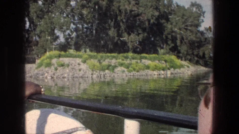 Sea of Galilee boat-view frame showing a rocky islet with grass and trees, the boat rail, a hand, and a partial passenger face at the edge.