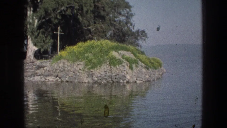 1976 8mm view from the Sea of Galilee showing a rocky green shoreline point with yellow flowers, trees, water, and a wooden cross.