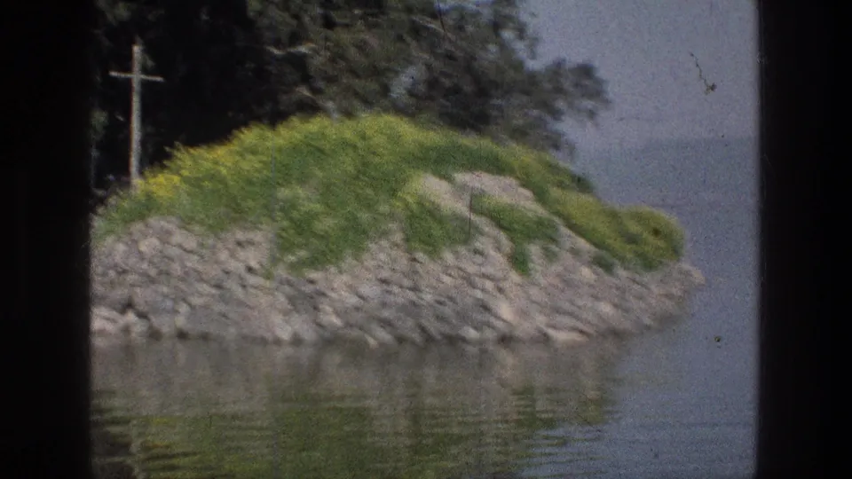 Sea of Galilee 1976 home-movie frame of a rocky, grass-covered shoreline point beside calm water, with trees and a wooden cross nearby.