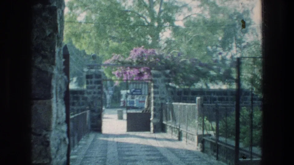 Handheld 8mm frame of the St. Peter's House approach, with stonework, railings, trees, and a small sign ahead.