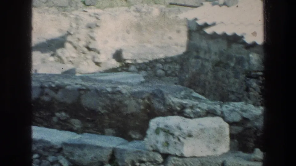 Handheld 8mm view of weathered stone remains at the Sea of Galilee site, with rubble and a small roofed structure beyond.