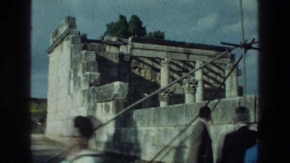1976 8mm frame near the Sea of Galilee showing tourists walking past ancient stone walls and metal supports.