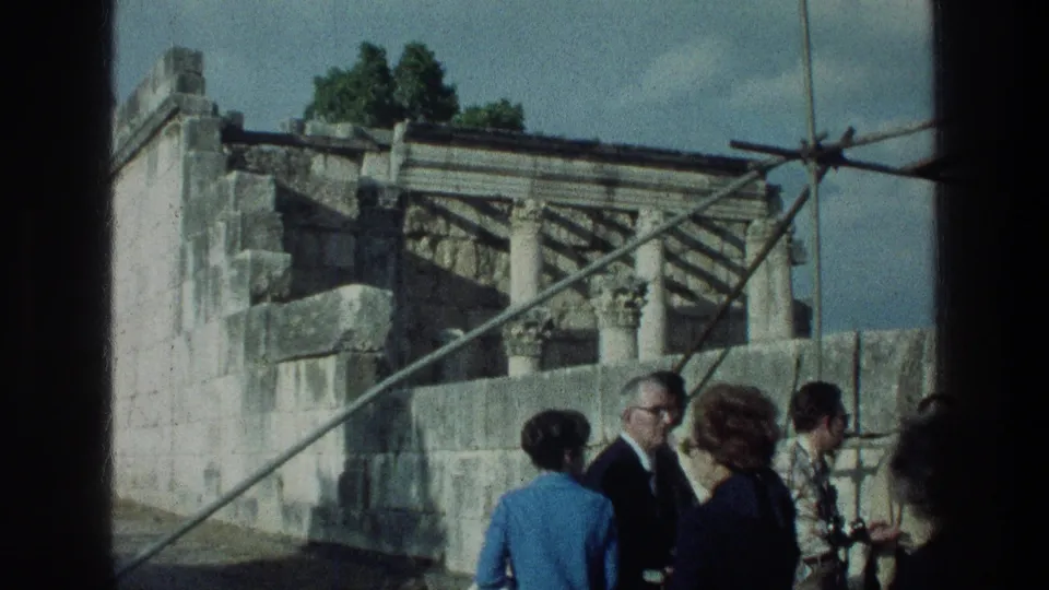 1976 home-movie frame of visitors near Sea of Galilee ruins, with suits, dresses, stonework and metal supports visible.