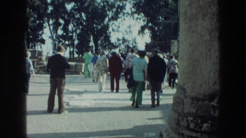 Tourists in period clothing walk deeper into the Sea of Galilee ruins, seen from behind under mottled outdoor light.