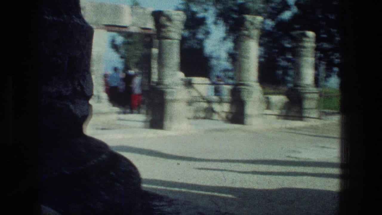 Tourists stand near columns and stone arch fragments at a sunlit ruin site.