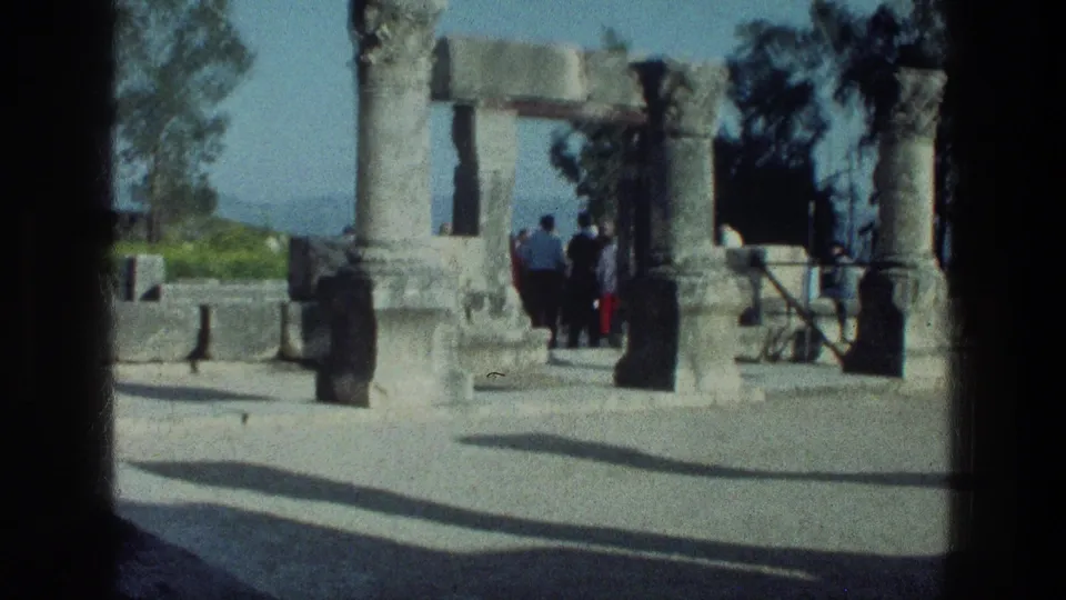 1976 8mm frame of visitors moving through ancient stone columns near the Sea of Galilee, with trees and hills beyond.