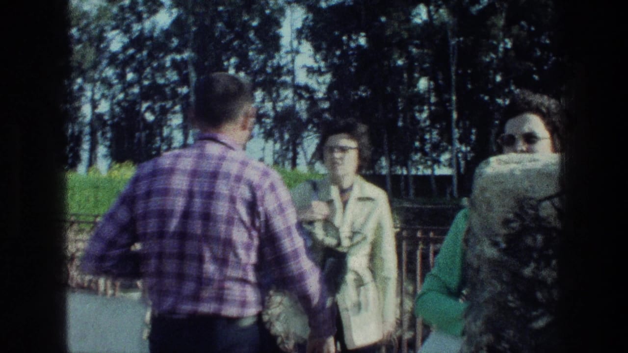 Adults converse outdoors near a railing and trees, with a man in a purple plaid shirt seen from behind.