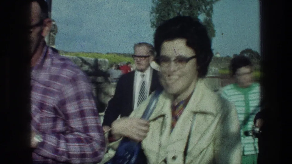 1976 Sea of Galilee home-movie frame with adults walking past the camera, including a woman in a cream coat and glasses.