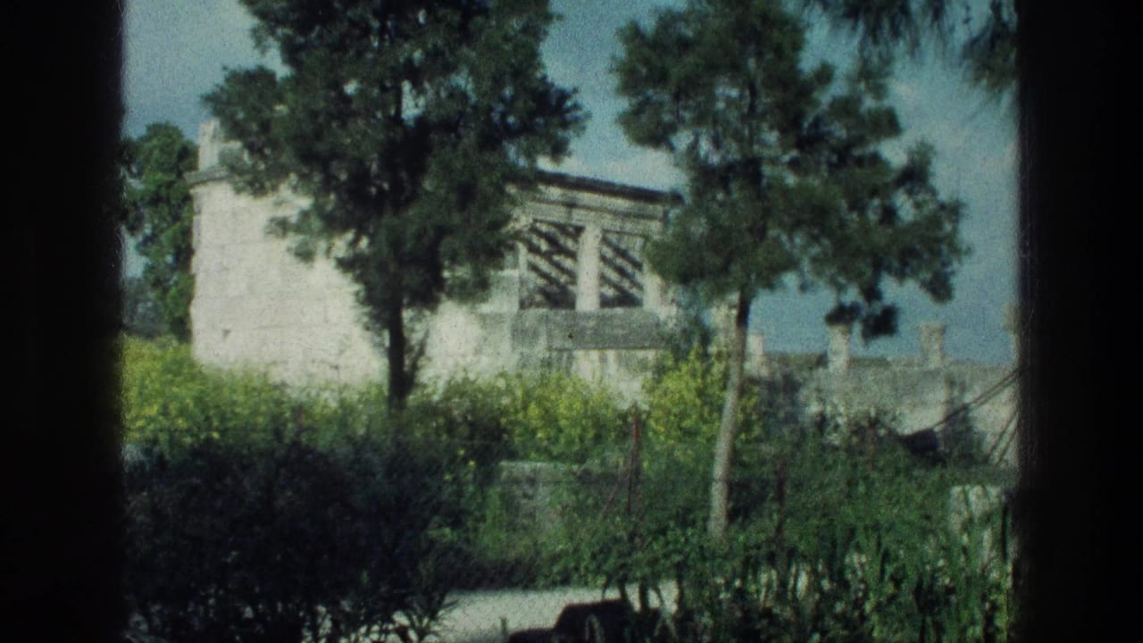 White stone building seen beyond trees and hedges through a dark vertical frame.