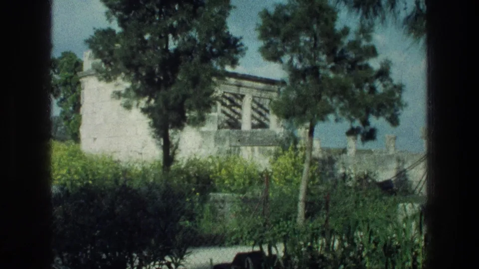 1976 Sea of Galilee home-movie frame showing a pale stone building partly hidden by trees, hedges, and a dark foreground frame.