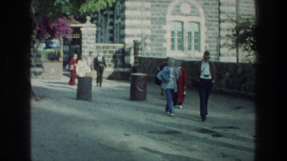 1976 Sea of Galilee frame showing pedestrians outside a gray stone building, with a gate, bins, trees, and paved walkway visible.
