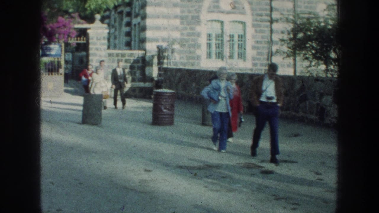 Pedestrians walk along a sunlit street beside a stone building and bins.