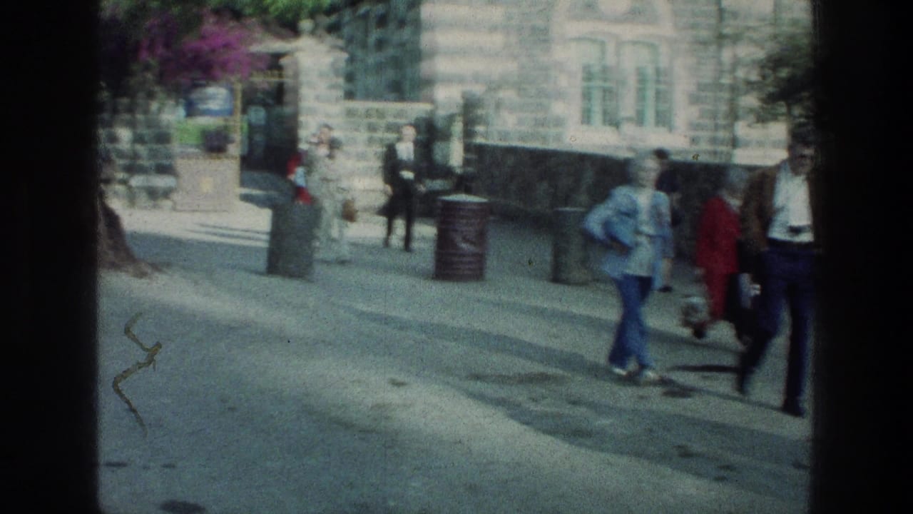 People in 1970s clothing move through a sunlit Sea of Galilee street scene with a stone building behind them.