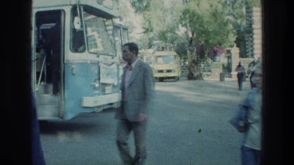 Blue and white bus with an open door as a pedestrian walks nearby on a sunlit 1976 Sea of Galilee street.