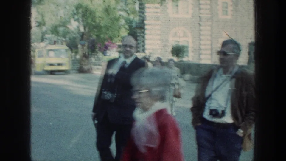 Older visitors carrying film cameras walk along a roadside near stone architecture during a 1976 Sea of Galilee stop.