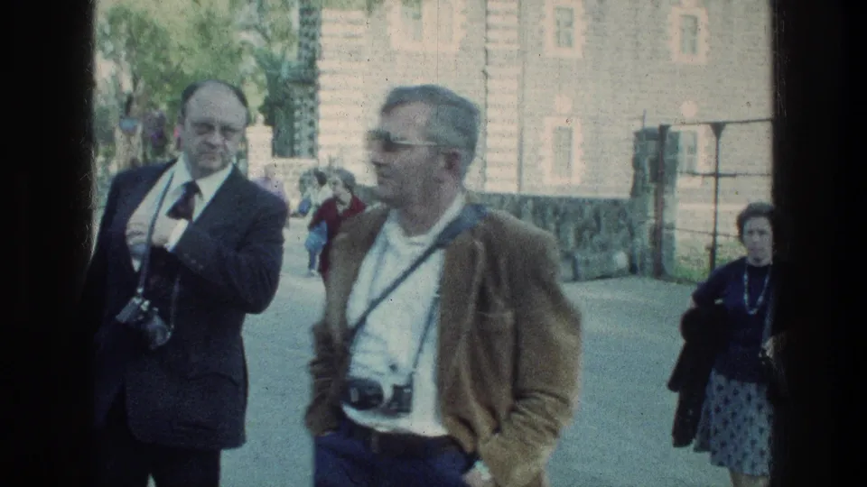 Two older male tourists with cameras stand near a roadside, with another visitor and stone building visible behind them.