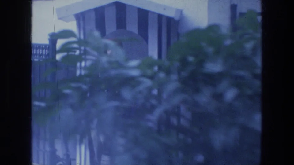 The guard post and iron railing appear behind foliage, with the uniformed figure partly hidden in the 1976 Tabgha footage.