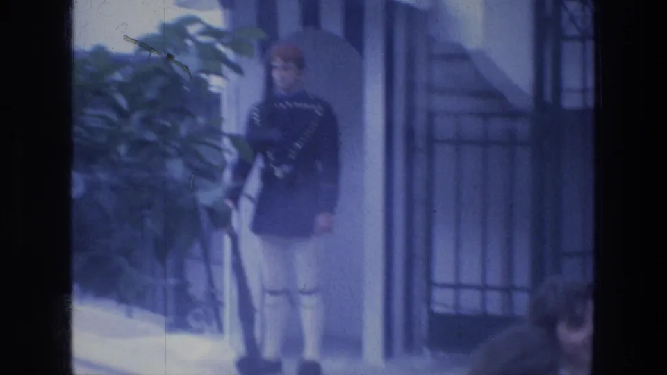 The guard stands in clearer view with a rifle beside foliage, a striped sentry post, and an iron gate in 1976 Tabgha footage.