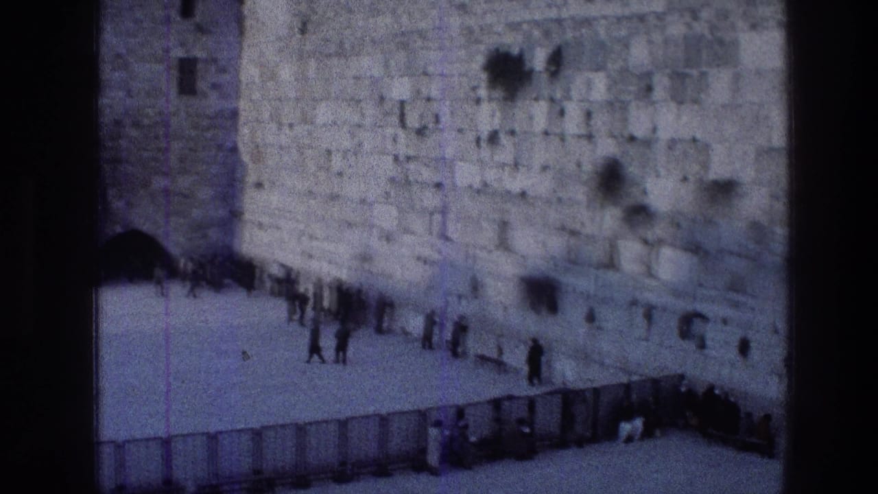 1976 8mm frame from Jerusalem showing the Western Wall courtyard from above, with small groups of people gathered near the limestone wall and metal barriers in the foreground.