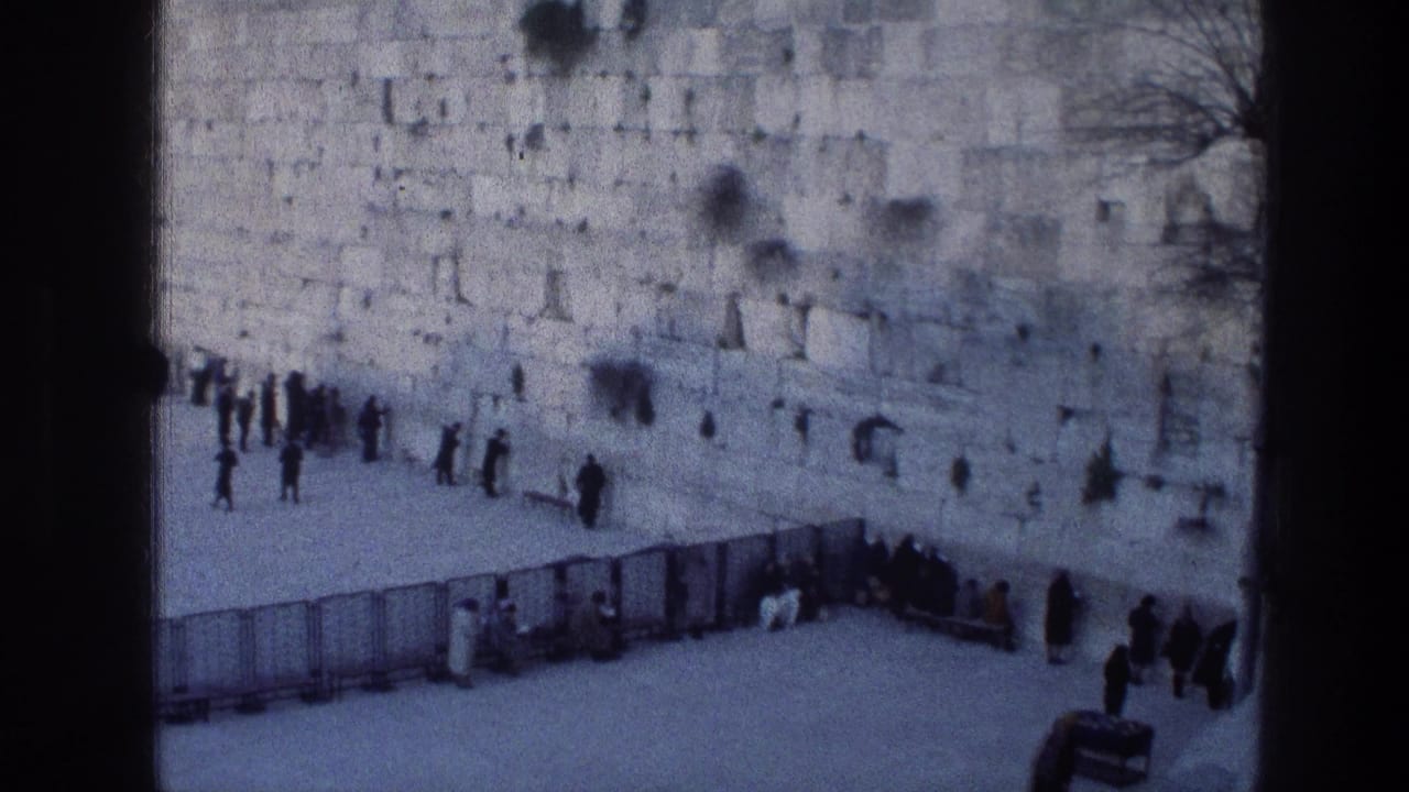 1976 8mm frame of the Western Wall in Jerusalem, viewed from an elevated position as people stand near the stone wall and move through the open courtyard.