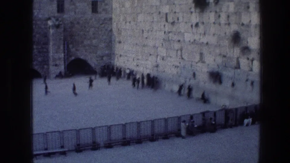 1976 8mm frame from the Western Wall courtyard, showing an arched opening at left, worshippers or visitors along the stone wall, and temporary barriers across the lower frame.
