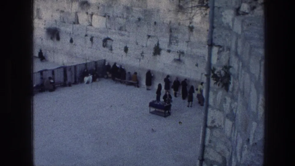 1976 8mm frame of the Wailing Wall courtyard with clustered worshippers, a small table and temporary barriers.