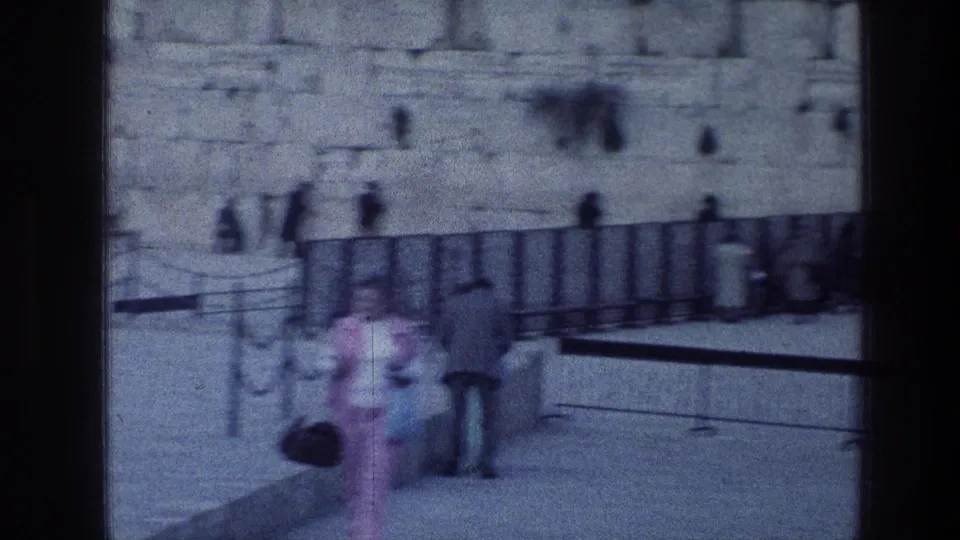 Blurred 8mm frame showing visitors, barriers, and the Western Wall area, with foreground figures partially obscuring the view.